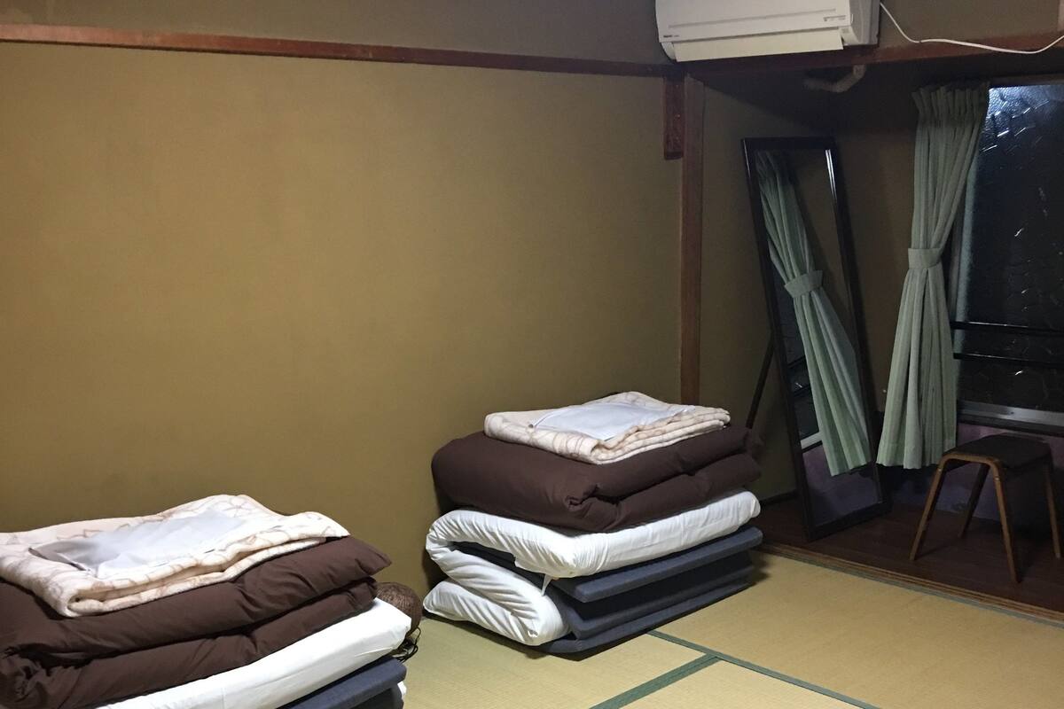 A room with tatami mats, two beds with folded bedding, an air conditioning unit, a mirror, and a window with curtains.