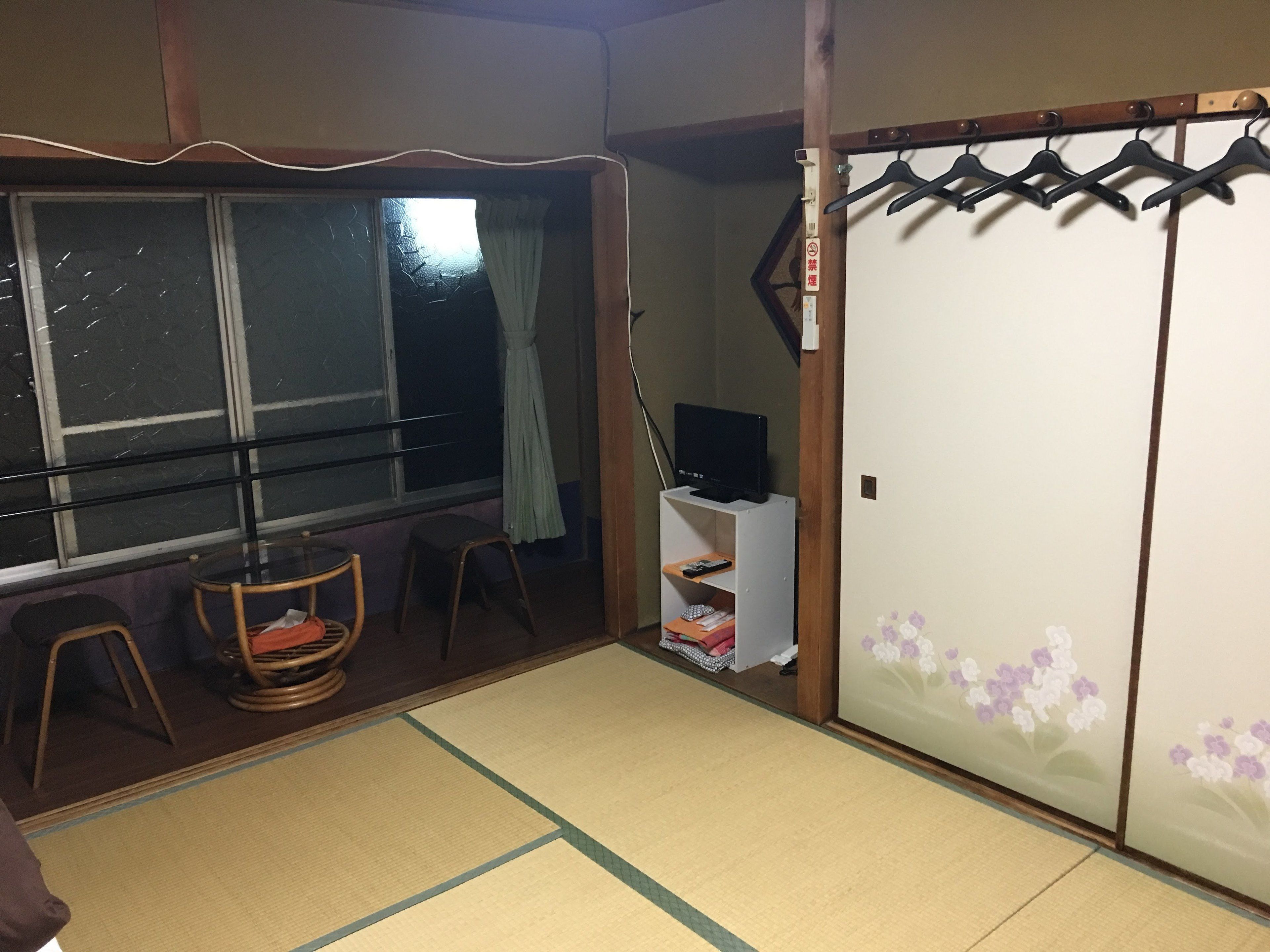 A traditional Japanese room with tatami flooring, a sliding door, a television on a stand, and a window with a curtain.