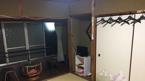 Ein traditioneller japanischer Raum mit Tatami-Bodenbelag, einer Schiebetür, einem Fernseher auf einem Standfuß und einem Fenster mit Vorhang.