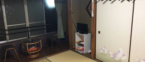 A traditional Japanese room with tatami flooring, a sliding door, a television on a stand, and a window with a curtain.