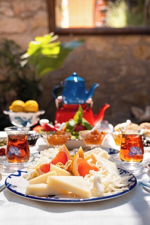 A table set with a plate of cheese, fruits, and tea cups, with a teapot and a bowl of lemons in the background.
