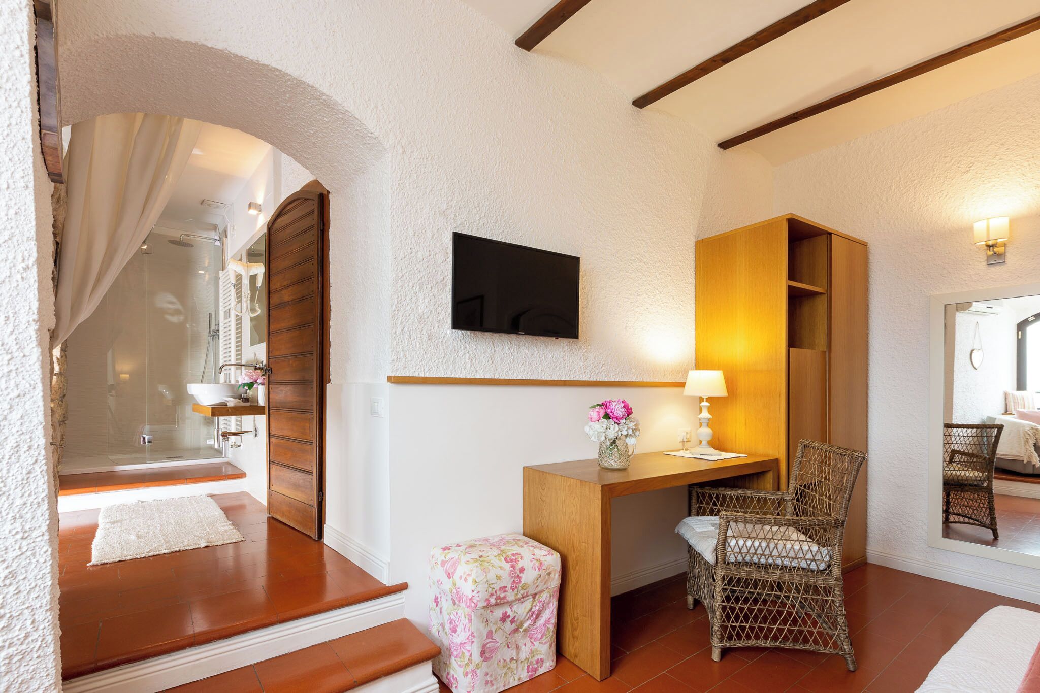 A hotel room with a wooden desk, wicker chairs, a TV mounted on the wall, a shower area, and a bed with a view of the room.