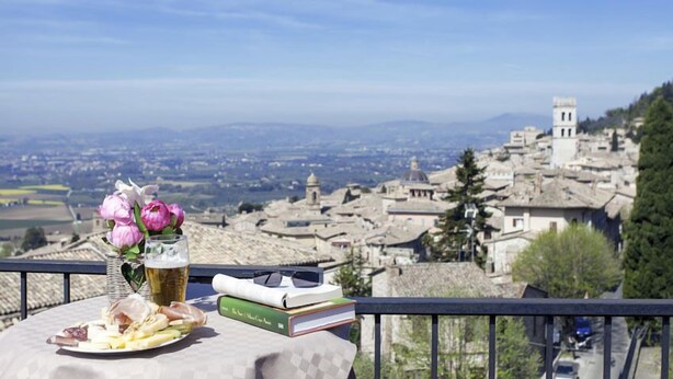A table with a plate of cheese, a glass of beer, and books, overlooking a historic town with a prominent bell tower. - Hotel Ideale (Assisi)