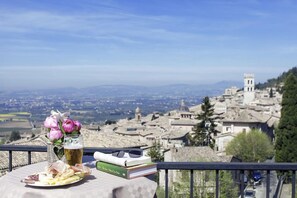 A table with a plate of cheese, a glass of beer, and books, overlooking a historic town with a prominent bell tower.