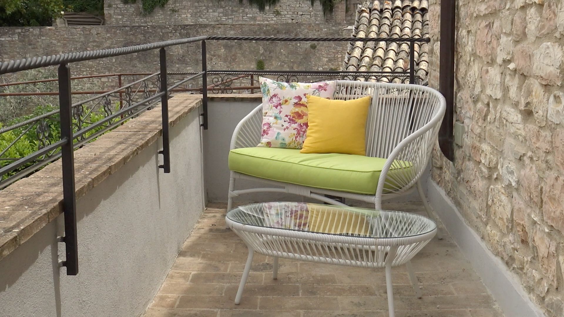 Un balcon avec une chaise en osier et des coussins, une rambarde en métal et un mur de pierre en arrière-plan.