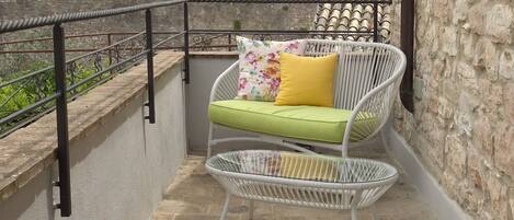Un balcon avec une chaise en osier et des coussins, une rambarde en métal et un mur de pierre en arrière-plan.