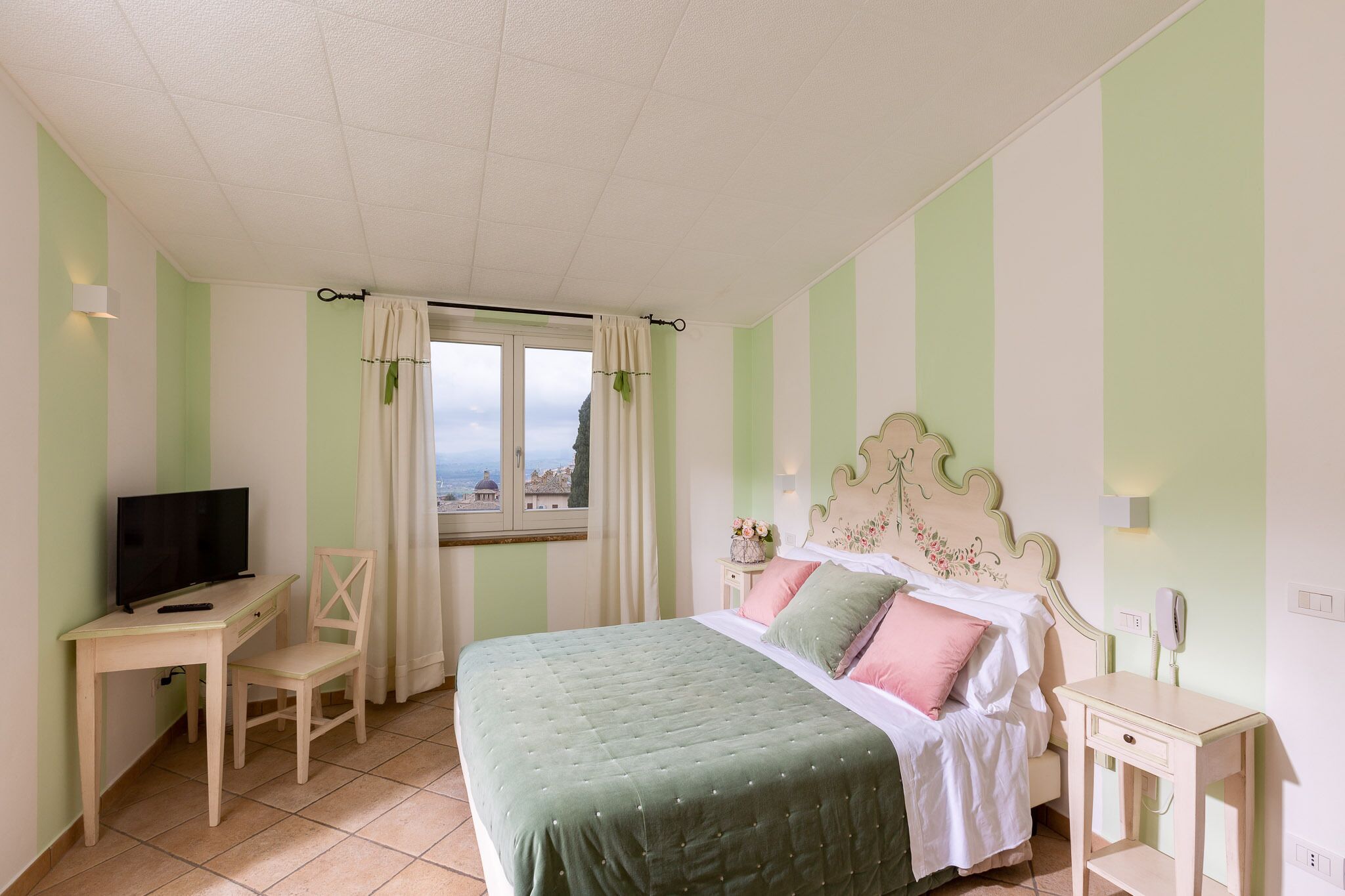 Une chambre à coucher comprenant un lit, un bureau, une chaise, un téléviseur et une fenêtre avec des rideaux.