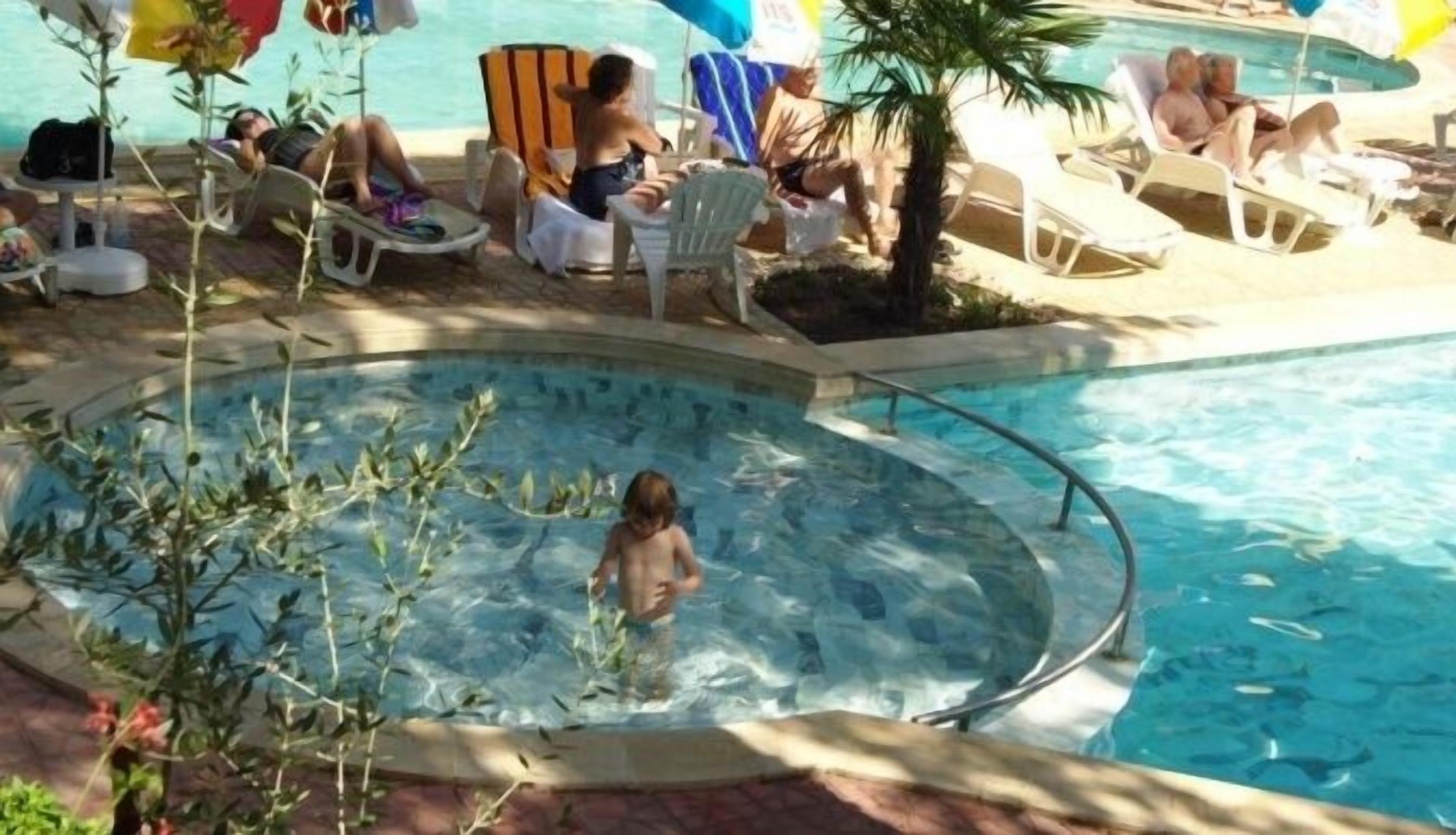 A pool area with lounge chairs and umbrellas, a child playing in the water, and a small garden area with plants.