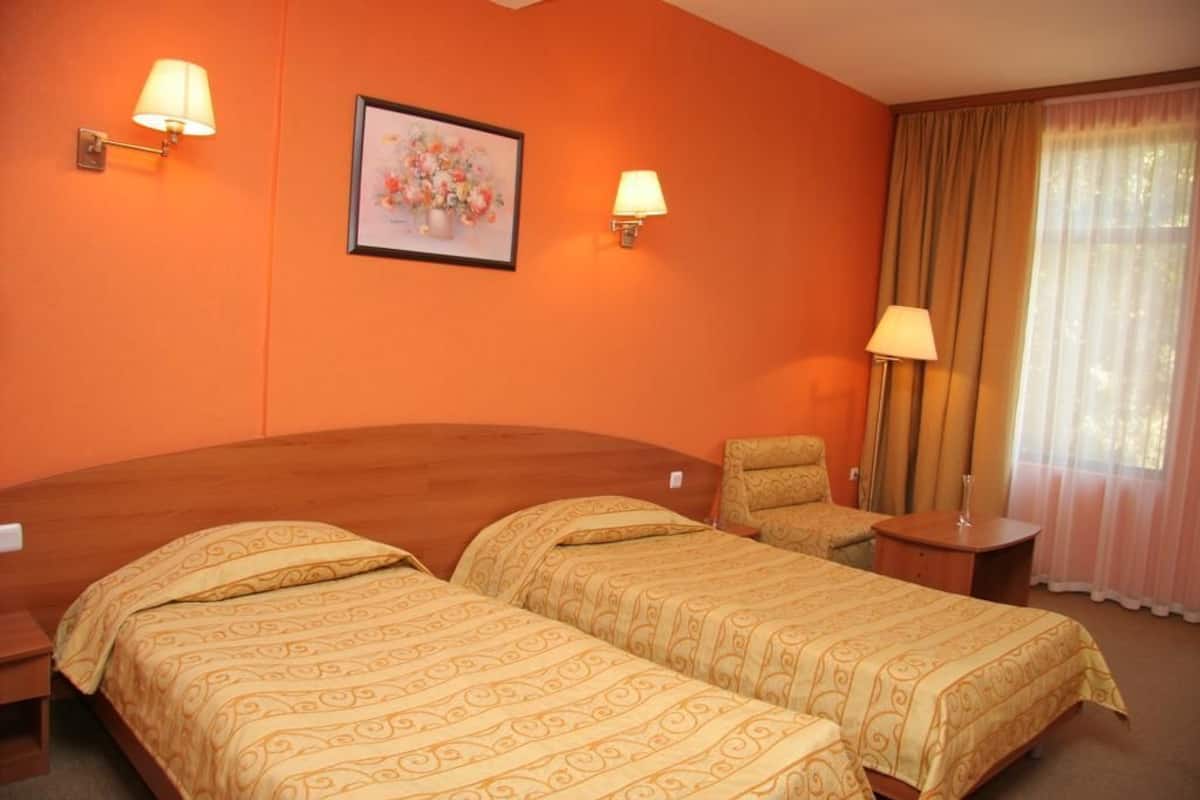 A hotel room with two single beds, a chair, a small table, and a framed floral artwork on the wall.