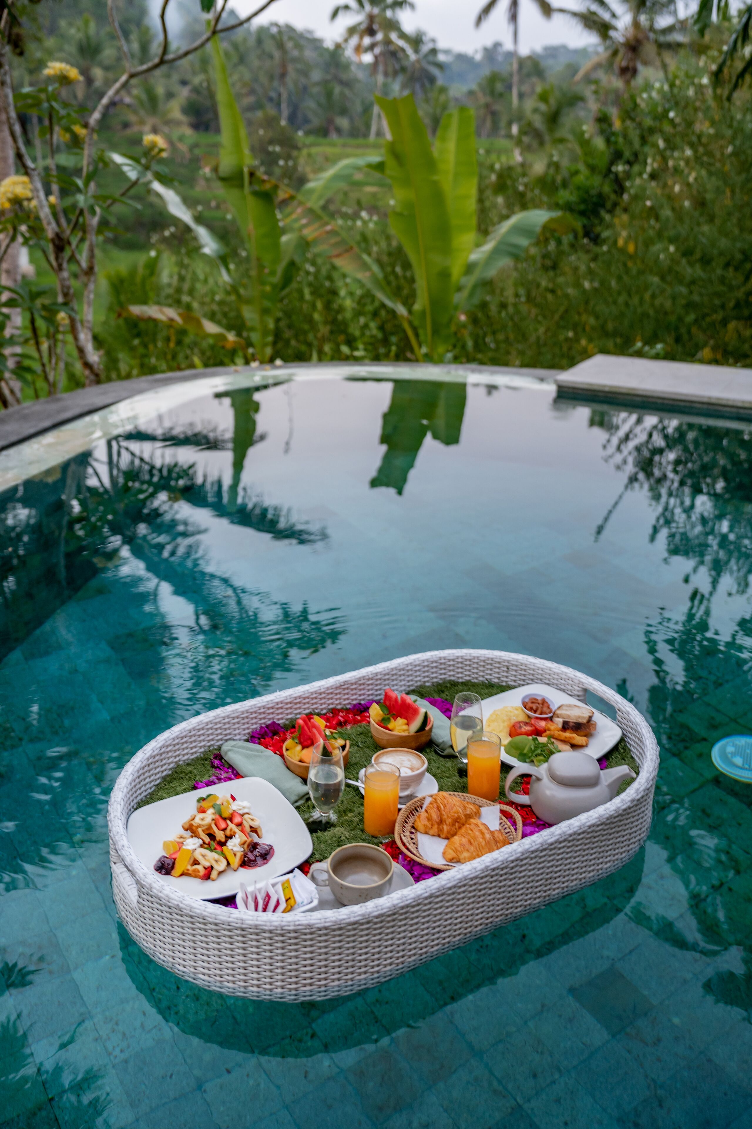 Bandeja de desayuno con comida y bebidas sobre una mesa de vidrio junto a una piscina.