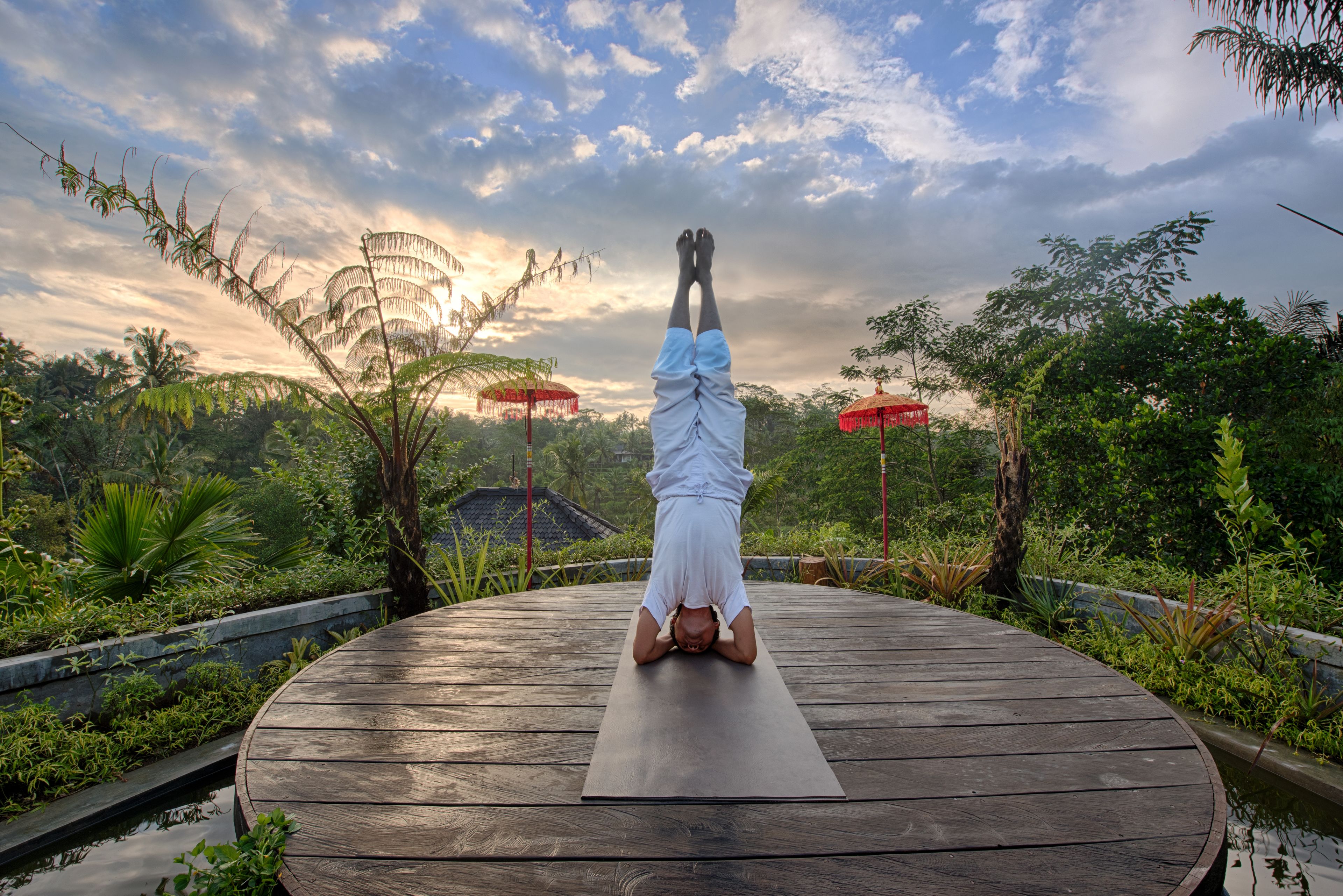 Une personne faisant la verticale sur une terrasse en bois, avec un tapis de yoga, entourée d’une végétation luxuriante et sous un ciel nuageux.