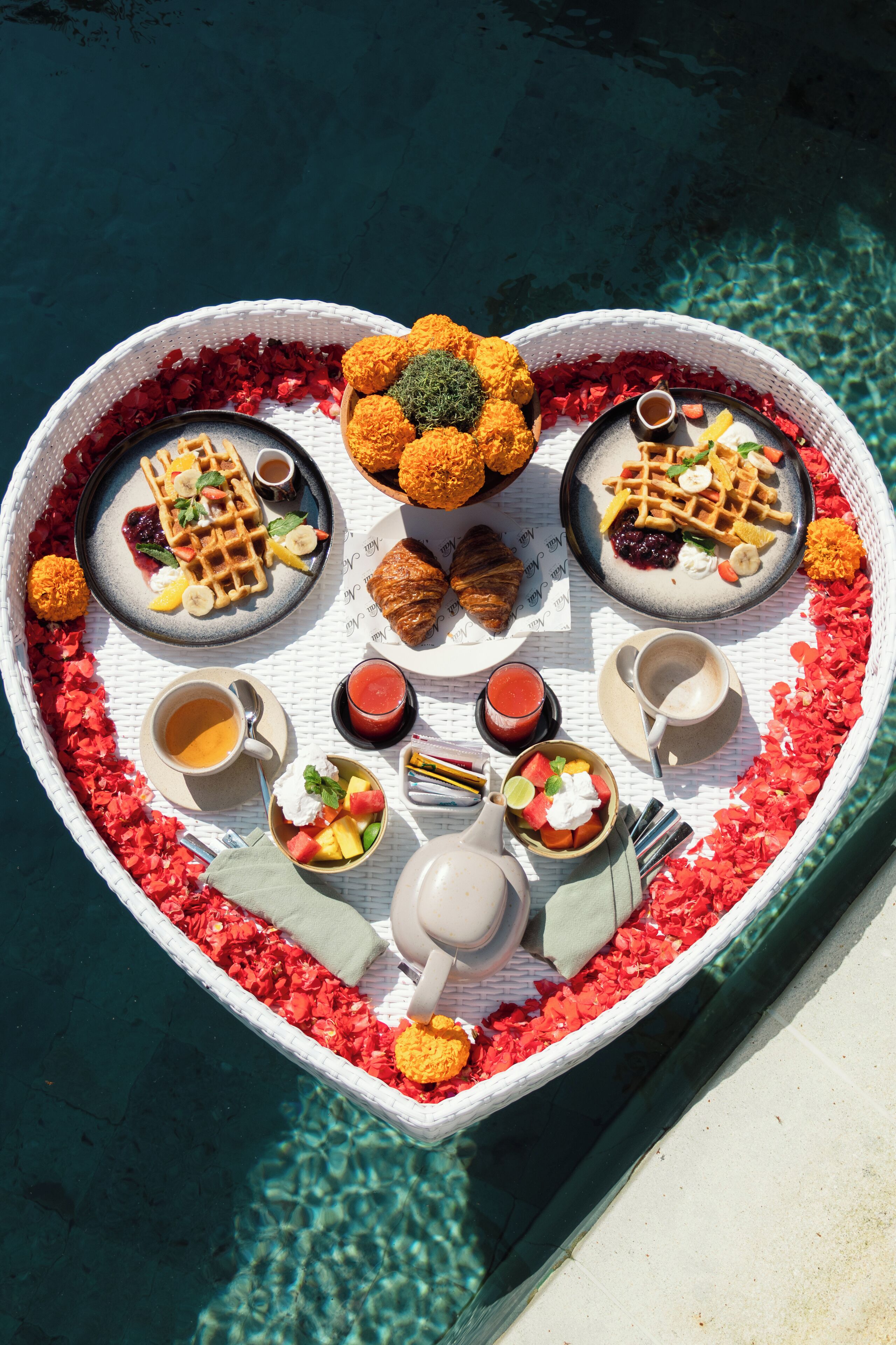 A heart-shaped tray filled with various breakfast items and flowers, placed on a poolside table.