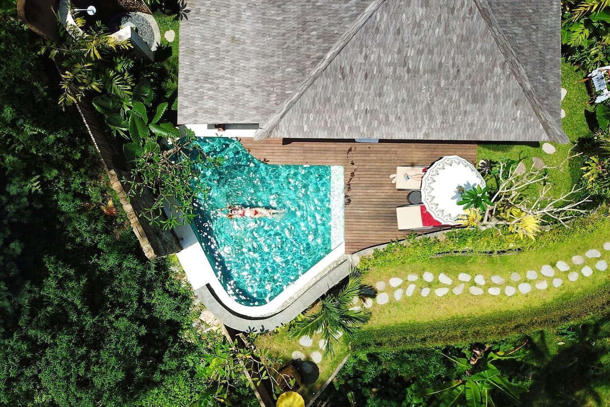 An aerial view of a house with a swimming pool, surrounded by lush greenery and a wooden deck.