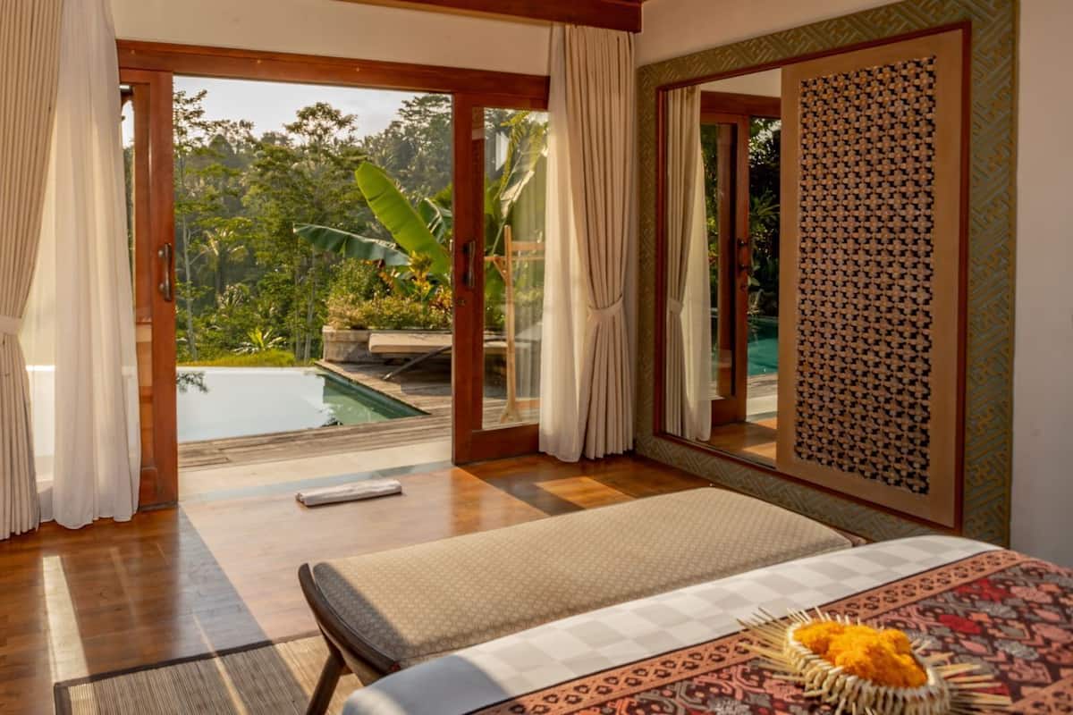 A room with a wooden floor, a bed with a patterned bedspread, a massage table, and a view of a swimming pool and greenery.