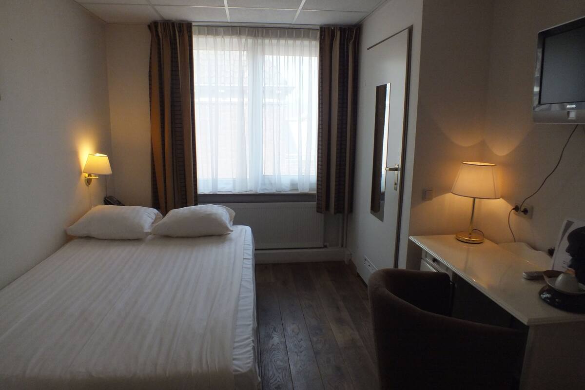 Ein Hotelzimmer mit Bett, Schreibtisch, Sessel, Fernseher und einem fenster mit Vorhängen.