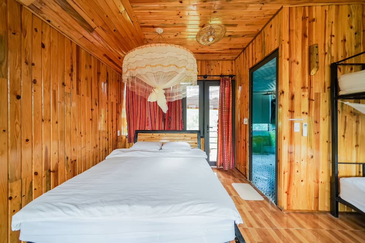 A wooden cabin room with a large bed, a hanging mosquito net, and a view through a glass door.