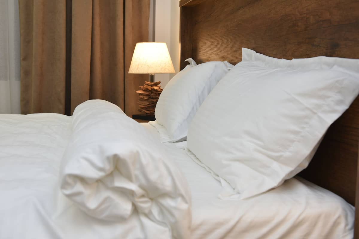 A neatly made bed with white linens and pillows, a wooden headboard, and a nightstand with a lamp and decorative item.