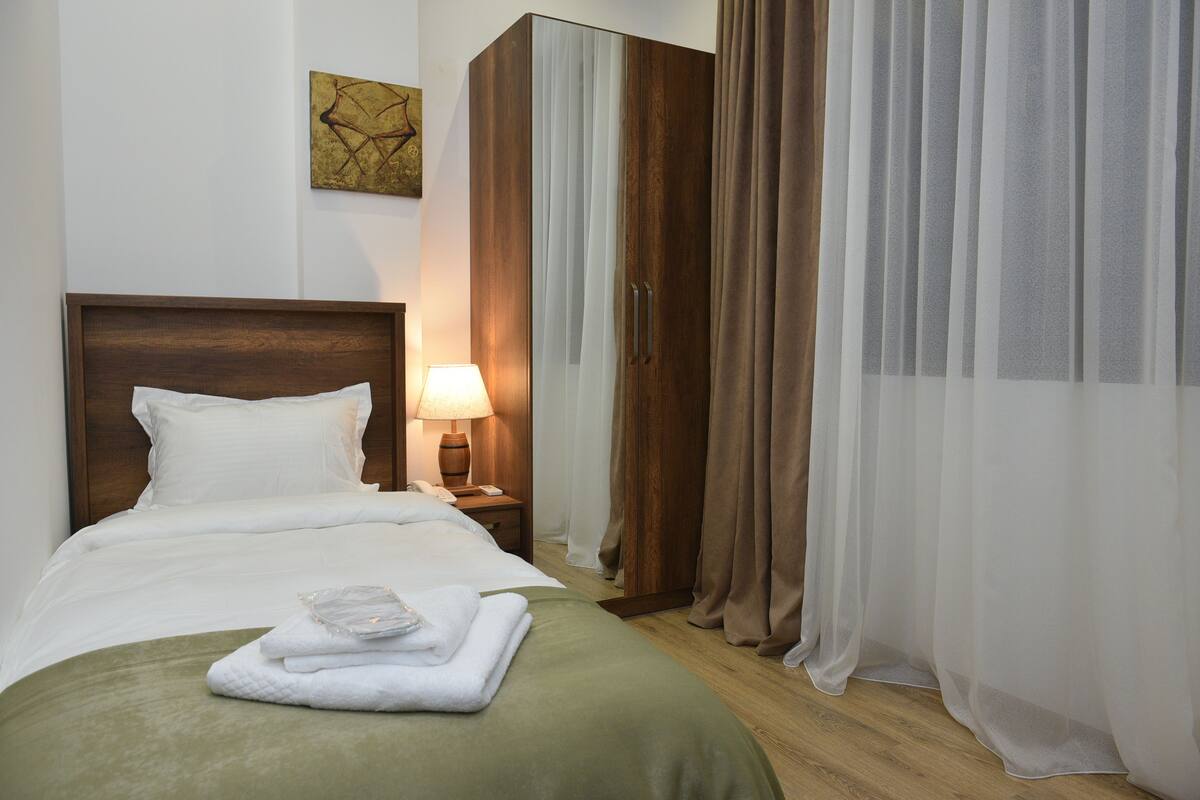 A neatly made bed with a green blanket, a wooden headboard, a bedside lamp, and a set of white towels.