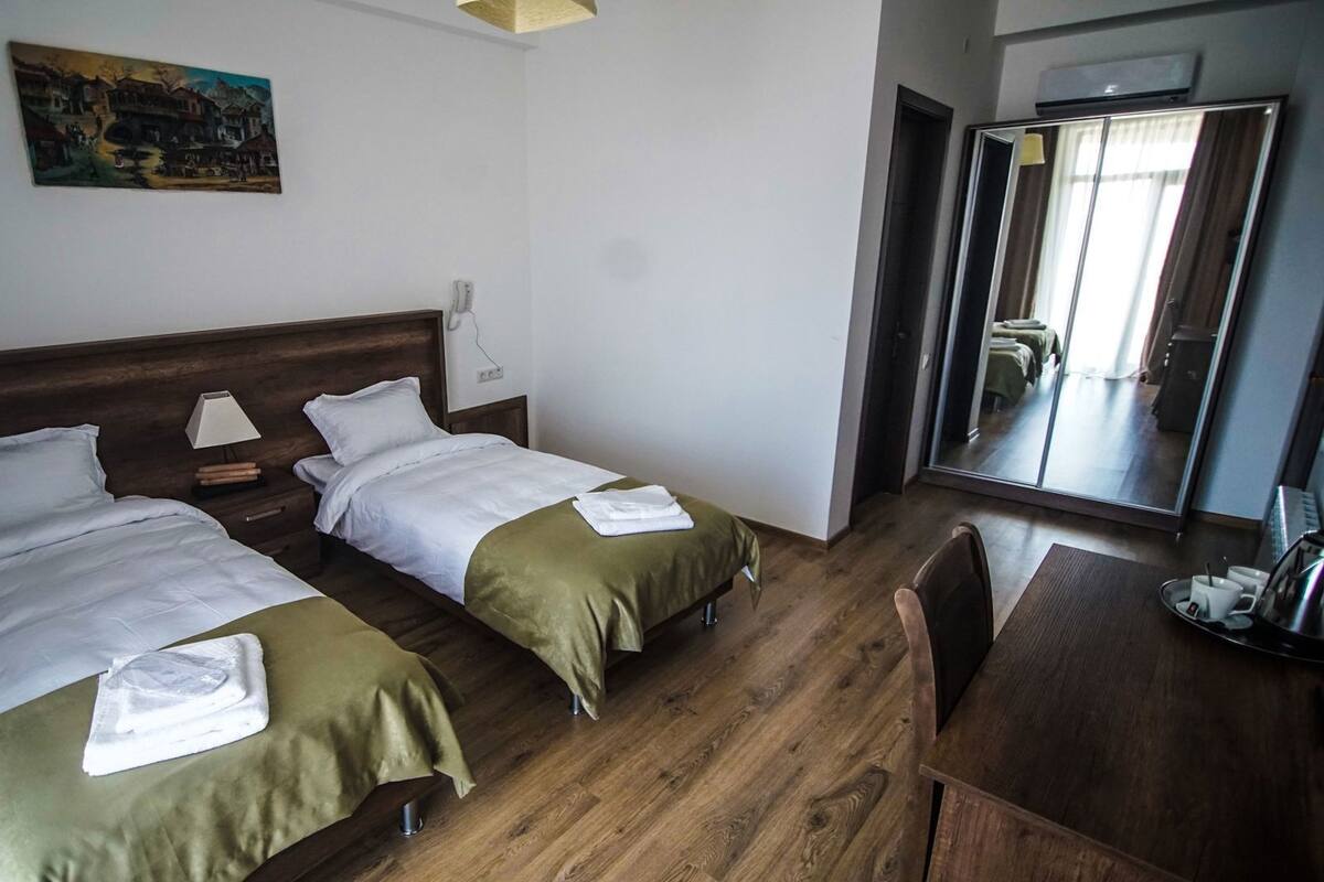 A hotel room with two beds, a desk, a chair, a painting on the wall, and a large mirror.