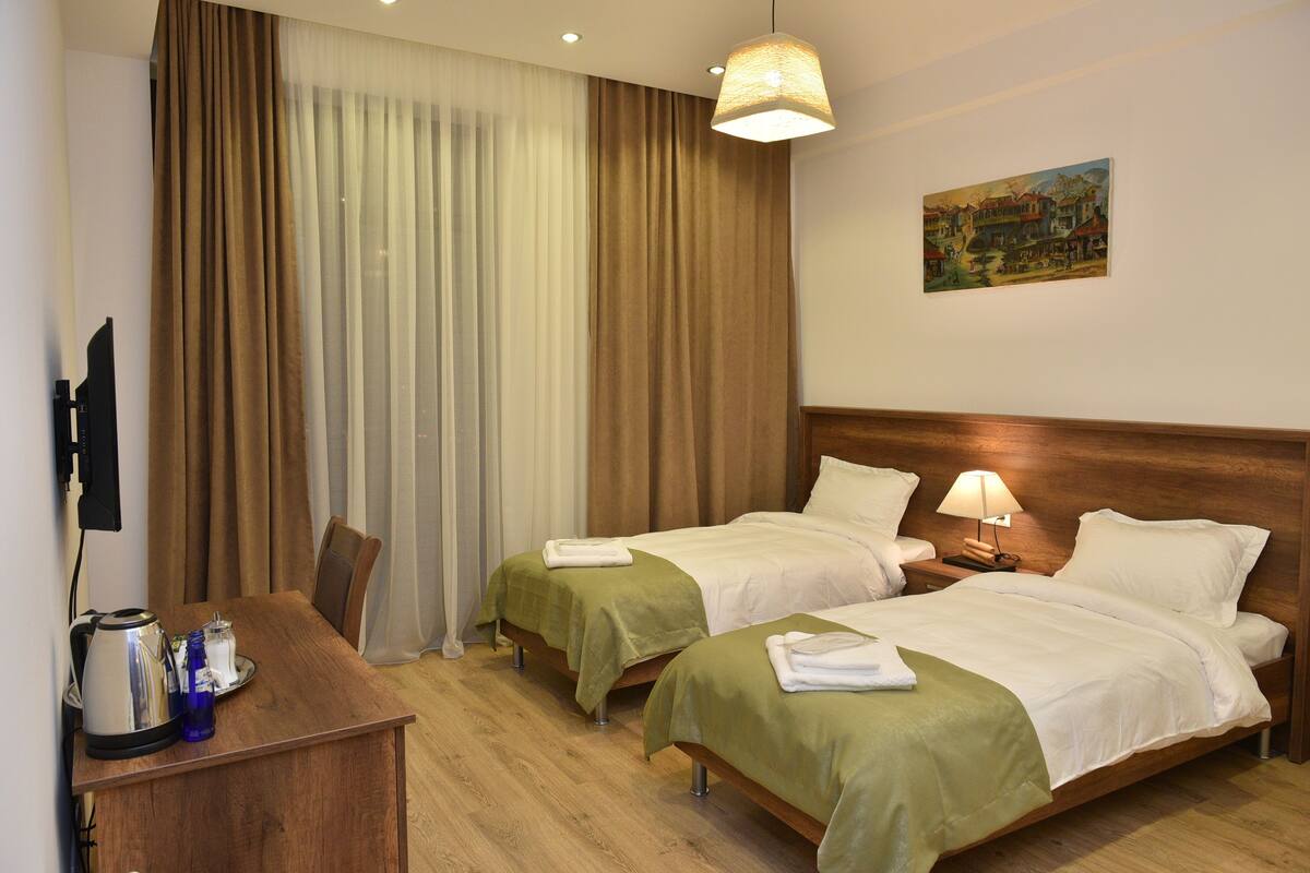 A hotel room with two beds, a desk with a kettle and a blue bottle, and a painting on the wall.