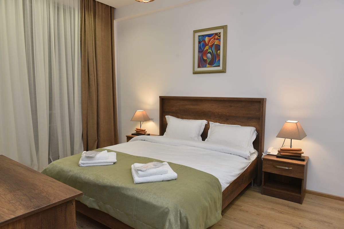 A neatly made bed with a green bedspread, two bedside lamps, a wooden nightstand, and a painting on the wall.