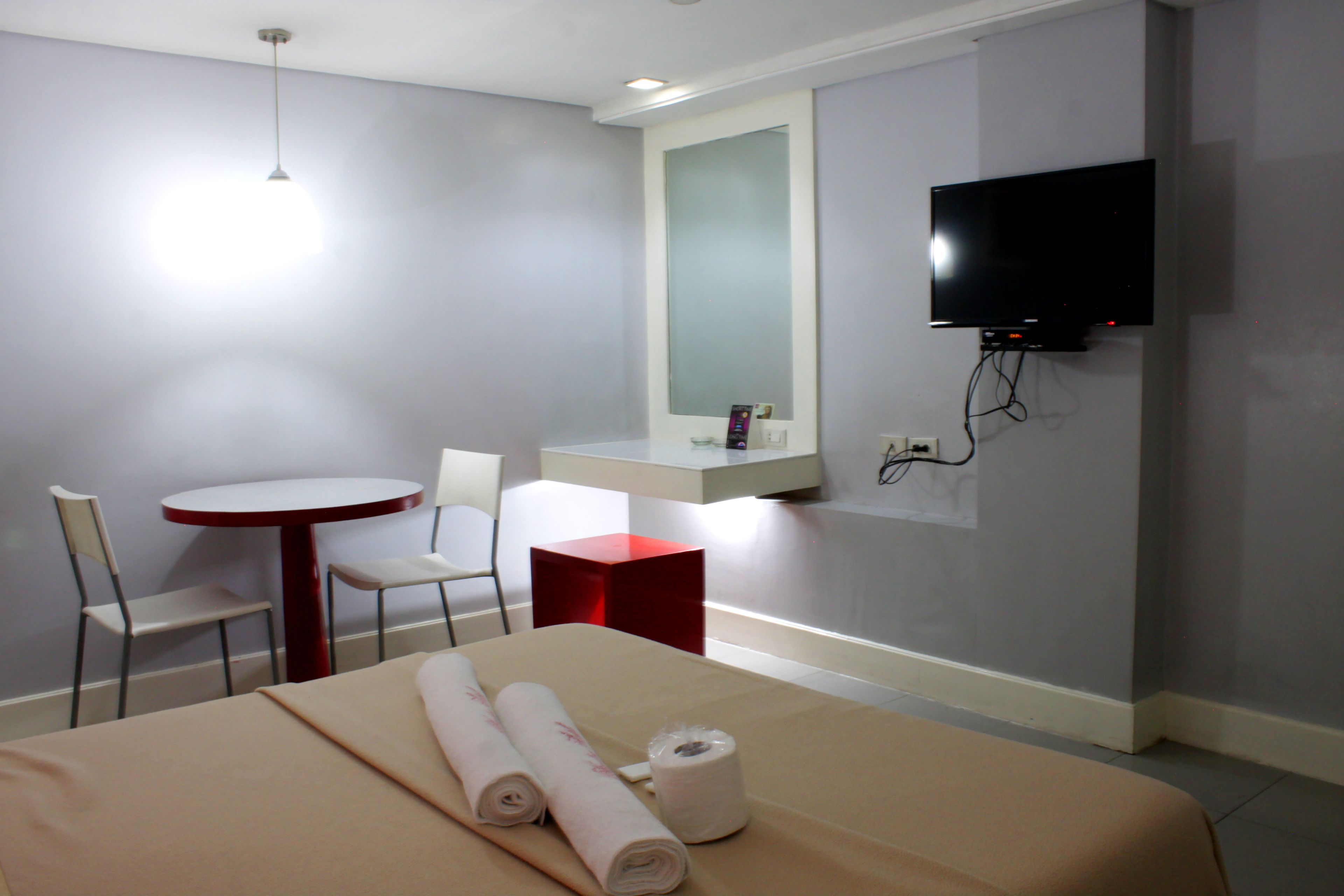 A hotel room with a bed, a round table, two chairs, a red cube-shaped object, a television mounted on the wall, and a ceiling light.