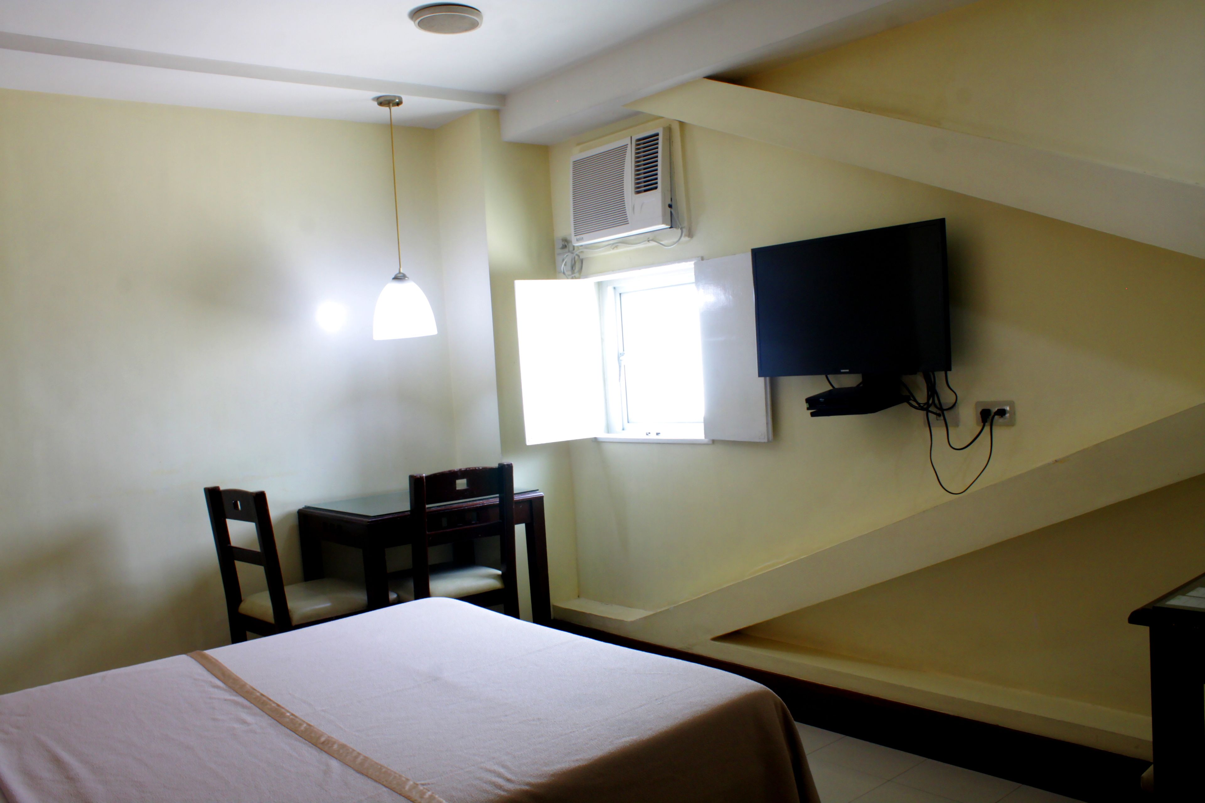 A hotel room with a bed, a desk, a chair, a television mounted on the wall, and an air conditioning unit.