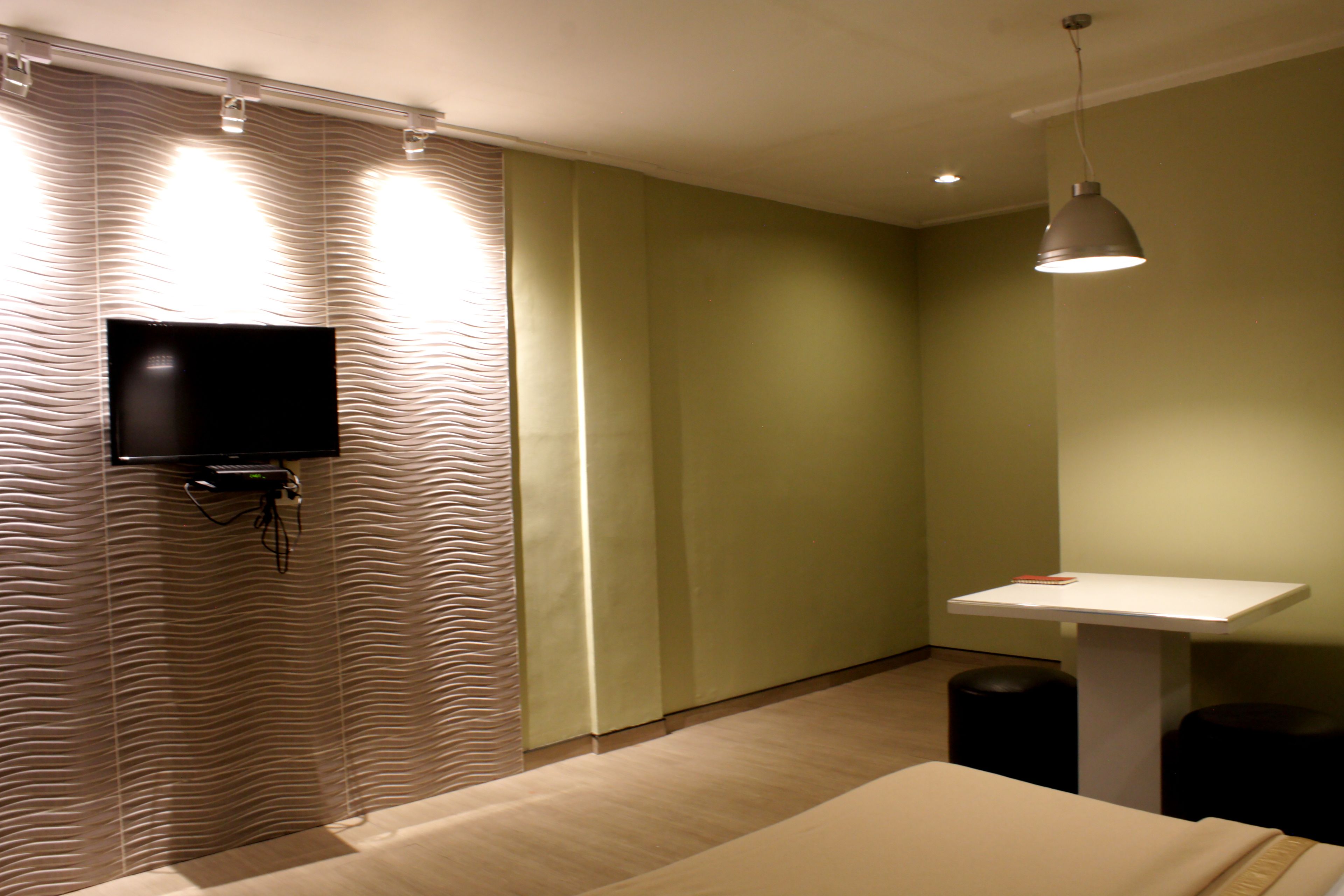 A hotel room with a bed, a television mounted on a wavy-patterned wall, a small table with a lamp, and two black stools.