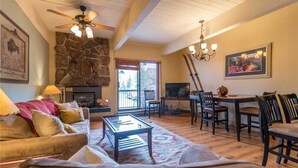Un salon chaleureux avec une cheminée en pierre, un ventilateur de plafond, un coin repas avec des chaises et un téléviseur.