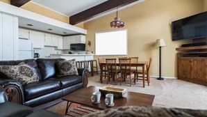 A living room with a black leather sofa, wooden dining table, and chairs. There is a TV mounted on the wall and a window with a view of the kitchen. - 410 Storm Meadows Club C Apartment 2 (Steamboat Springs)