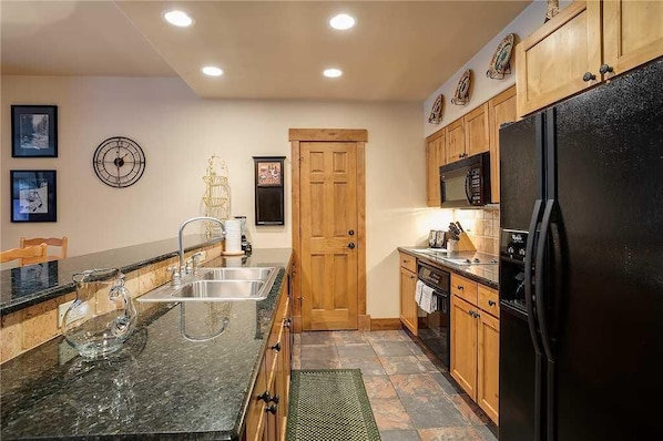A kitchen with a granite countertop, stainless steel sink, wooden cabinets, a black refrigerator, and a microwave.