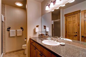 A bathroom with a granite countertop, a white sink, and a large mirror. There are two towels hanging on hooks and a wooden door in the background.
