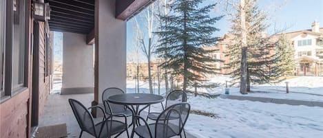 A covered patio with a table and chairs, a wooden exterior wall, and a view of snow-covered ground and trees.