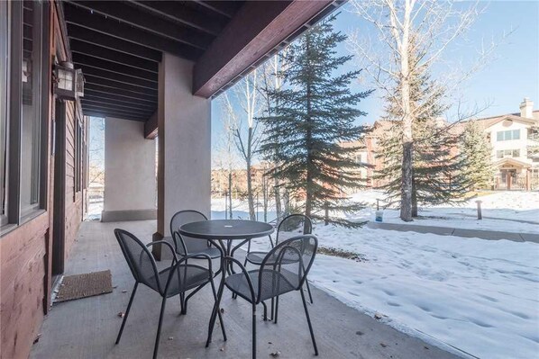 A covered patio with a table and chairs, a wooden exterior wall, and a view of snow-covered ground and trees.