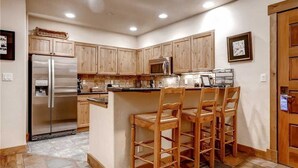 A kitchen with wooden cabinets, a stainless steel refrigerator, a microwave, and a bar area with stools. - 3109 Champagne Lodge, Trappeur's Crossing Apartment 3 (Steamboat Springs)