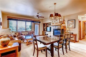 A spacious living room with a large wooden dining table, comfortable seating, a television, and a kitchen area with a stone backsplash. - 3110 Champagne Lodge, Trappeur's Crossing Apartment 3 (Steamboat Springs)