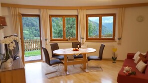 A room with a wooden dining table, chairs, a red sofa, and large windows with a view of a forested landscape.