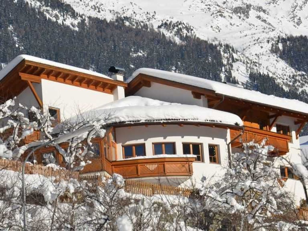 Un edificio de estilo chalé con balcón de madera y techo cubierto de nieve, rodeado de árboles y montañas nevadas.