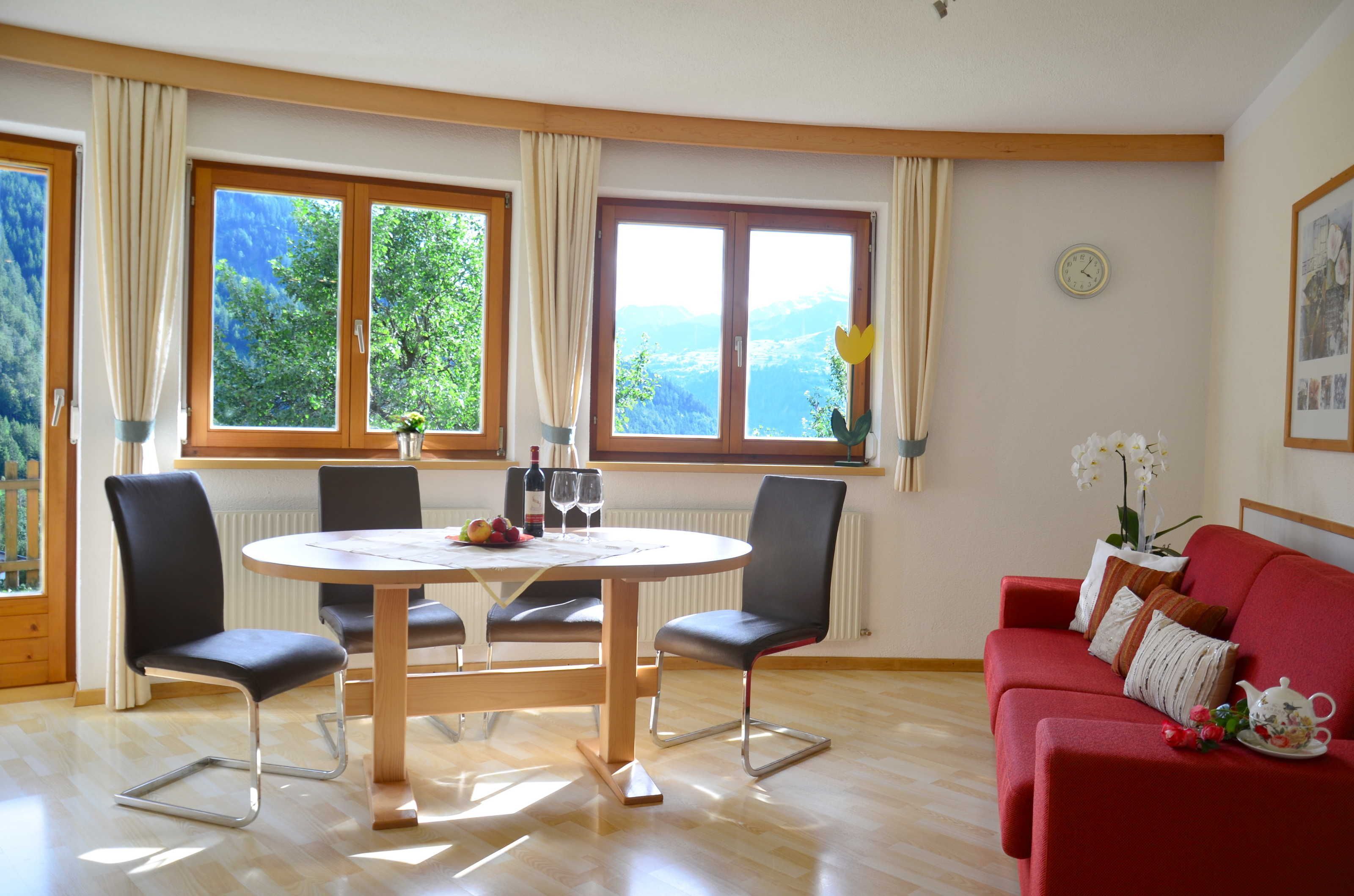 A room with a dining table, chairs, a red sofa, and large windows with curtains.