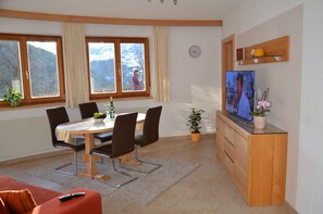 A dining area with a table, chairs, a TV, a clock, and a window with a view of mountains.