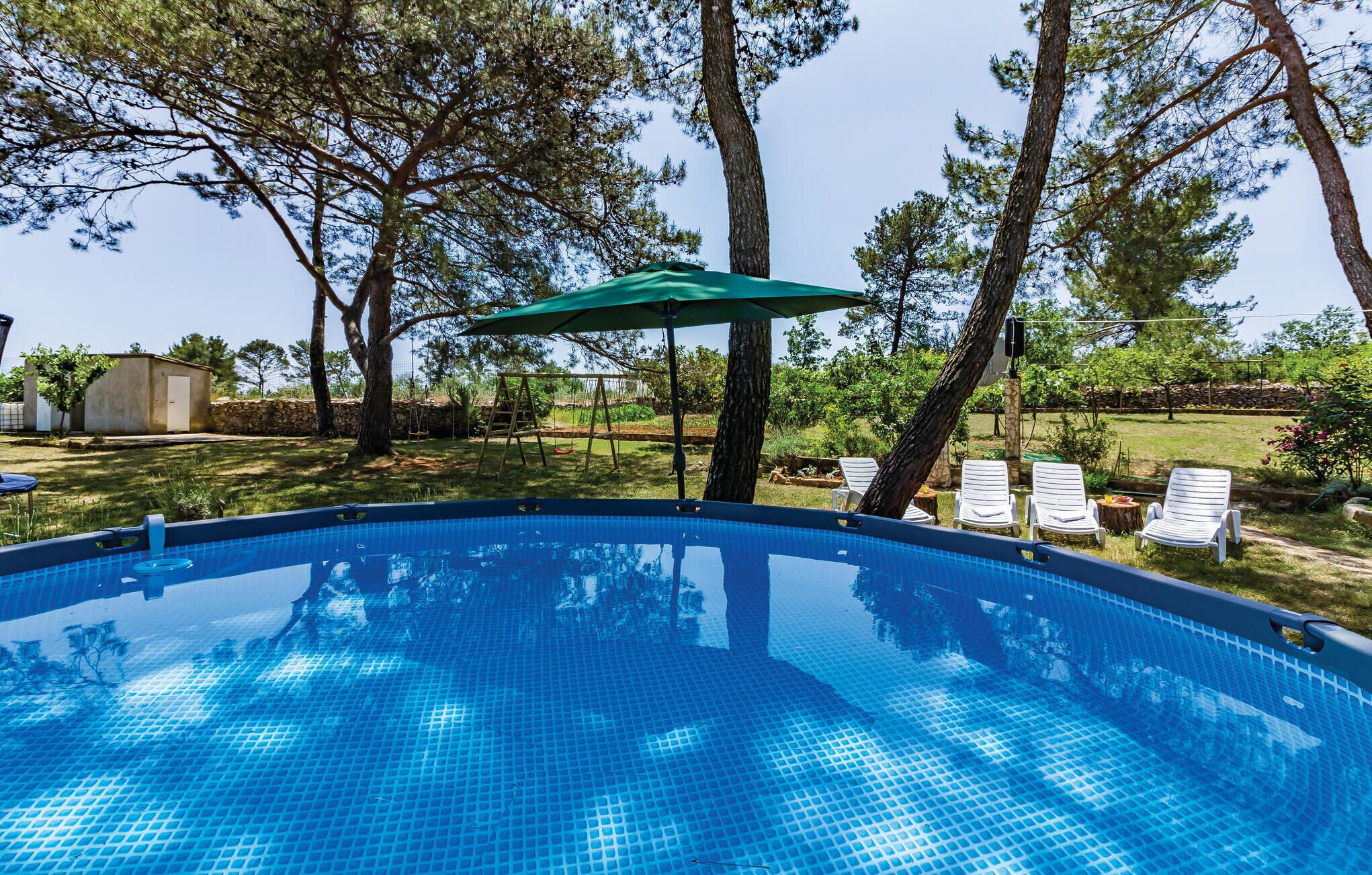 Piscina com guarda-sol verde, cercada por árvores e mobiliário de exterior.