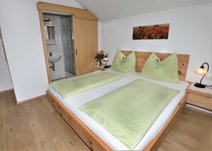 Un dormitorio con cabecera de cama de madera, dos almohadas, una mesita de noche con una lámpara, otra mesita más pequeña con un jarrón de flores, y un área de ducha con puerta de vidrio.