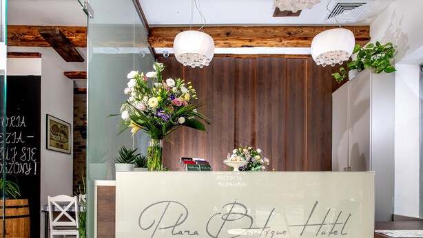 Un comptoir d’accueil agrémenté d’une composition florale, une enseigne indiquant « Plaza Boutique Hotel », et un plafond en bois.