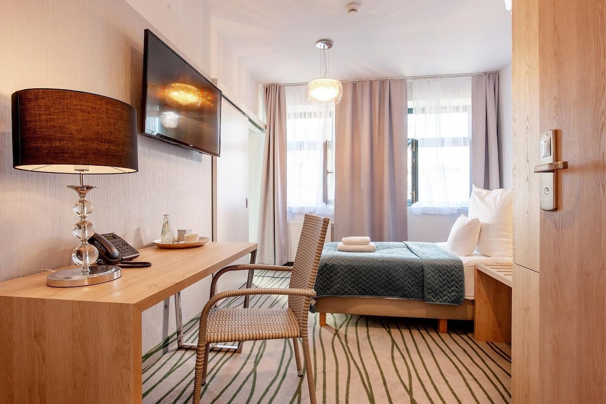 A hotel room with a bed, a desk with a telephone, a chair, a television mounted on the wall, and a window with curtains.