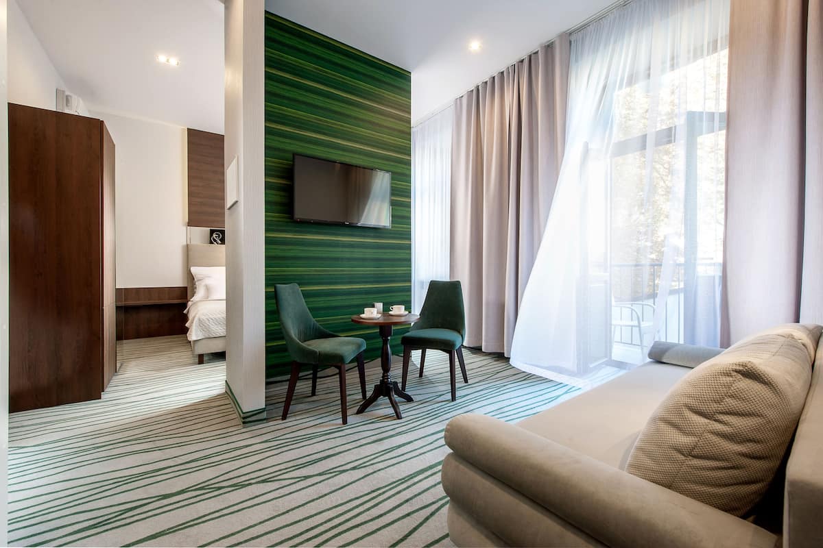 Une chambre d’hôtel moderne avec un mur d’accent vert, un téléviseur à écran plat, un canapé, deux fauteuils, une petite table avec des tasses et une vue sur le balcon.