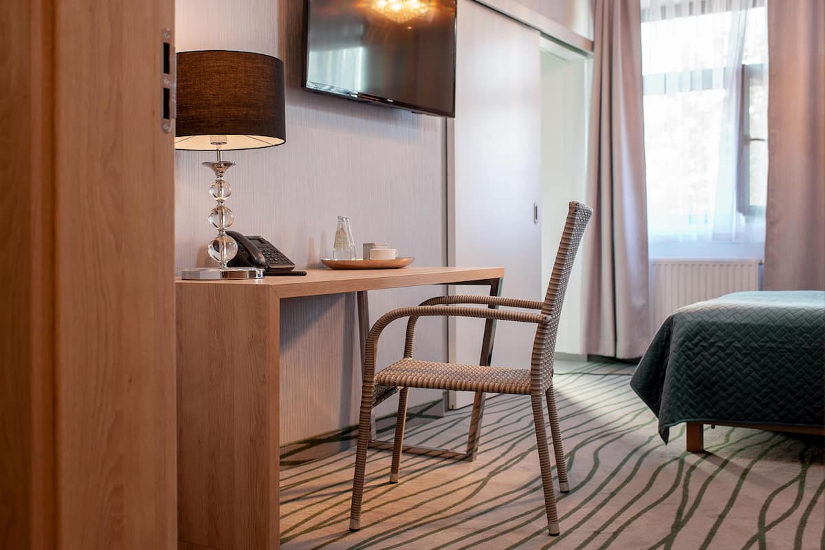 Une chambre d’hôtel avec un bureau en bois, une chaise en osier, un lit recouvert d’une couette verte, un téléviseur fixé au mur et une fenêtre avec des rideaux.