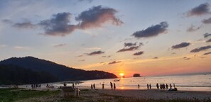 A beach scene with a sunset, people on the sand, and boats in the water.