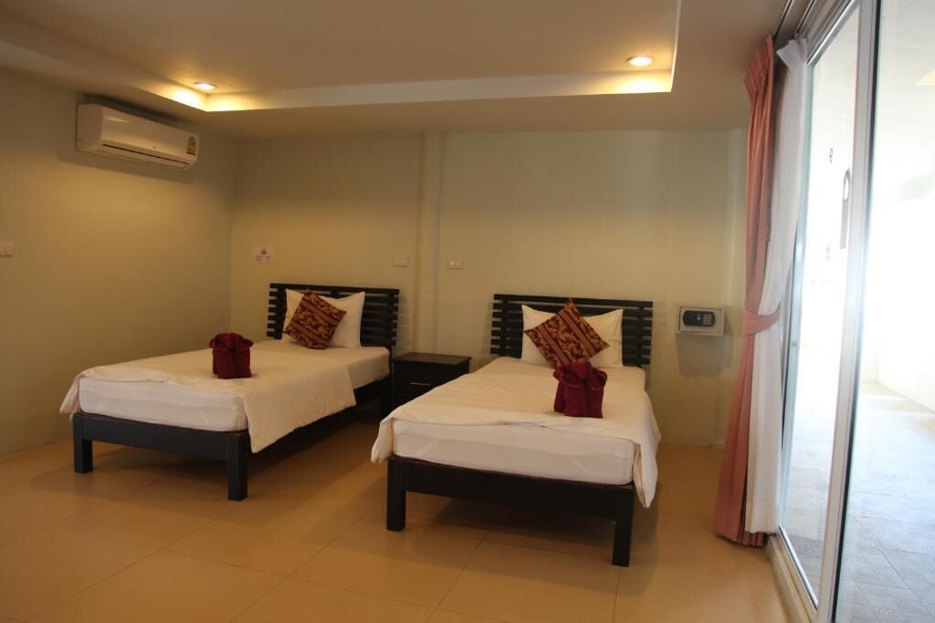 A hotel room with two single beds, each with a pillow and a red item on the bed. There is a window with curtains, an air conditioning unit on the wall, and a small table between the beds.