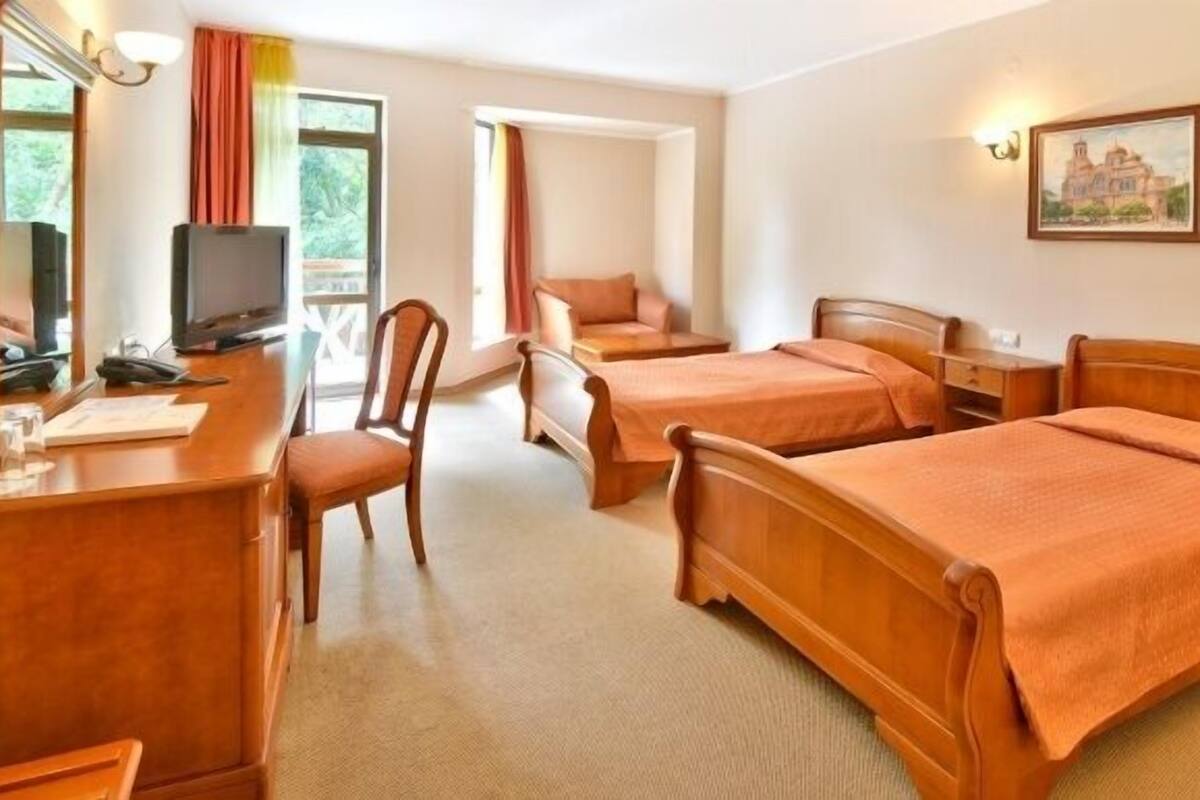 A hotel room with two single beds, a desk with a chair, a television, and a window with curtains.