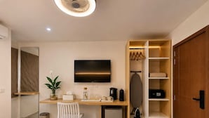A hotel room with a bed, a desk with a computer, a TV, a small fridge, a plant, and a wardrobe.